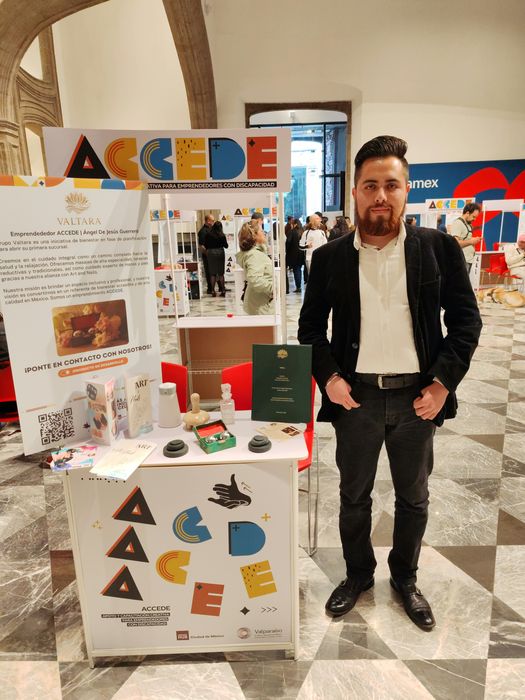 Ángel de Jesús Guerrero Vizzuett, de pie con saco negro y camisa blanca, junto al estand de Valtara con el letrero ACCEDE, iniciativa para emprendedores con discapacidad. Detrás, el cartel de Valtara con su descripción, código QR y la leyenda ¡Producto en desarrollo!; en la mesa, folletos de Art & Nails, piedras para masaje, esferas chinas en su estuche y un proyecto encuadernado en verde. Al fondo, otros estands en un patio de arcos de piedra.