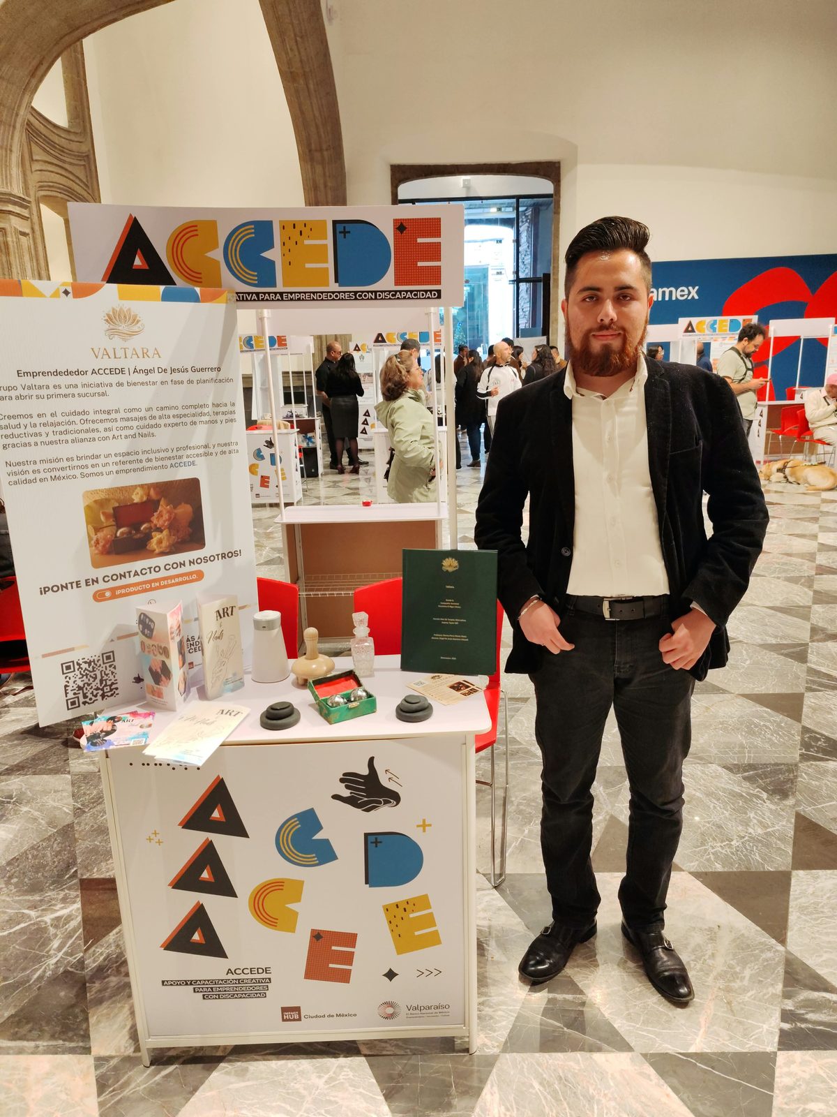 Ángel de Jesús Guerrero Vizzuett, de pie con saco negro y camisa blanca, junto al estand de Valtara con el letrero ACCEDE, iniciativa para emprendedores con discapacidad. Detrás, el cartel de Valtara con su descripción, código QR y la leyenda ¡Producto en desarrollo!; en la mesa, folletos de Art & Nails, piedras para masaje, esferas chinas en su estuche y un proyecto encuadernado en verde. Al fondo, otros estands en un patio de arcos de piedra.