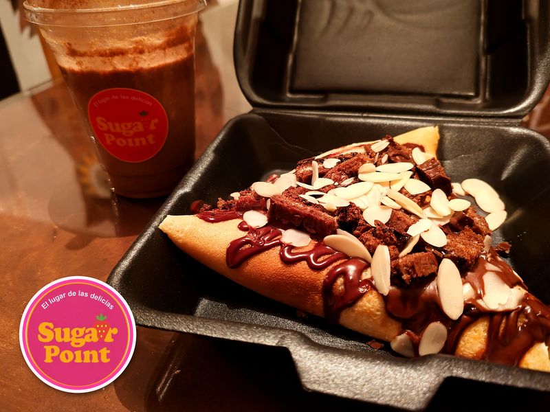 Crepa doblada en triángulo, servida en un contenedor negro, bañada con hilos de chocolate y cubierta con trozos de pastel de chocolate y almendras fileteadas; atrás, un vaso con bebida de chocolate y canela con la etiqueta roja de Sugar Point. En la esquina, el logotipo de Sugar Point.